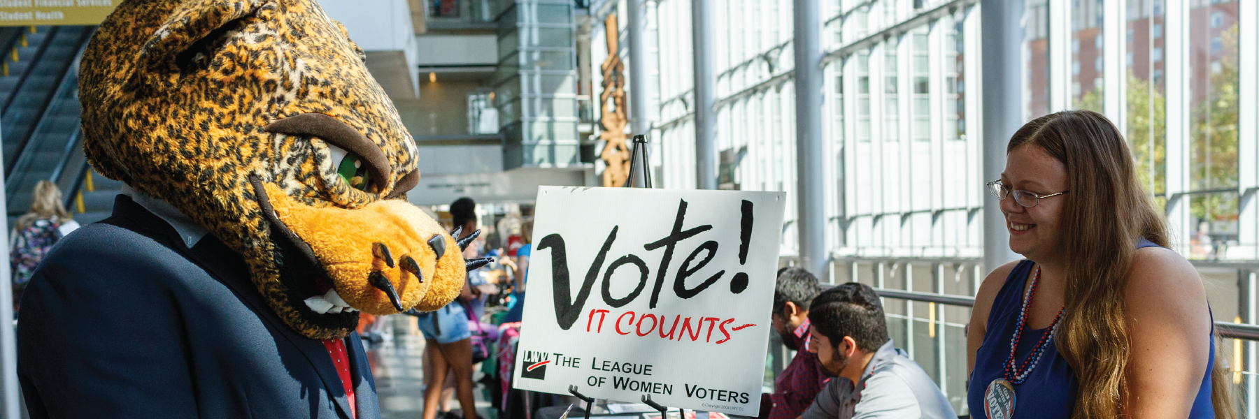 A person wearing a jaguar head and a suit looks at a woman in front of a sign that says vote.