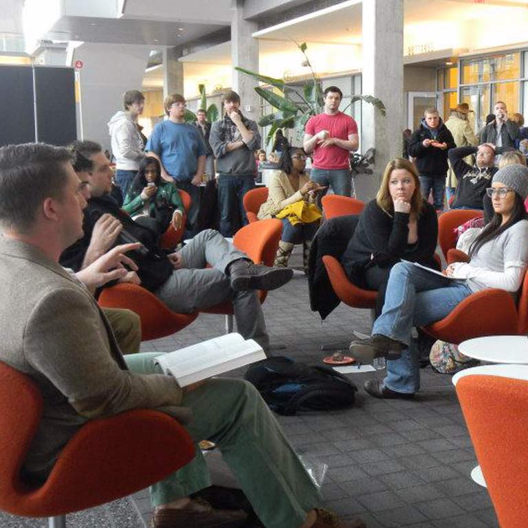 A group of people sitting in orange chairs. Another group stands behind them.